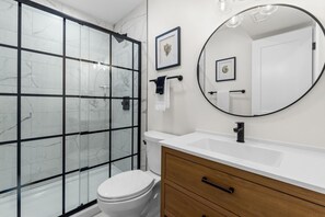 Un baño con cabina de ducha de vidrio, inodoro blanco, tocador de madera con lavamanos y espejo.