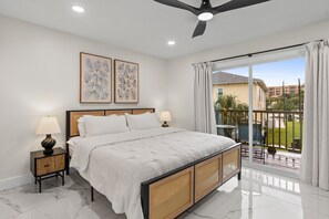 Un dormitorio con una cama, dos mesitas de noche, un ventilador de techo y vistas a un balcón con mobiliario exterior.