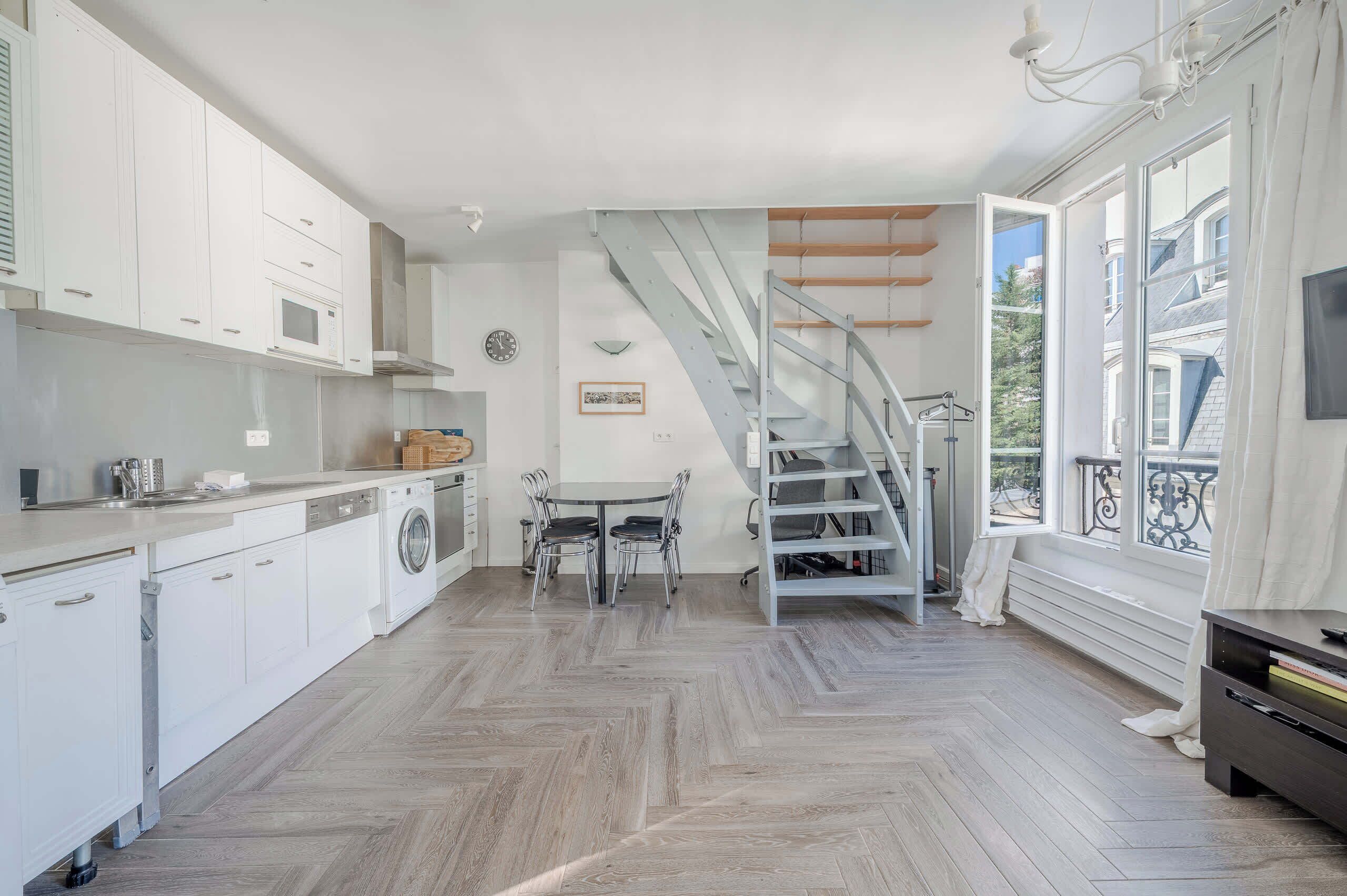 A modern kitchen with white cabinetry, a built-in washing machine, a dining area with a table and chairs, and a staircase leading to an upper level.