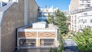 Um edifício de vários andares com fachada de pedra, uma varanda com grades de metal e uma pequena área de jardim com árvores e arbustos.