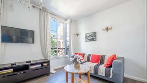 Uma sala de estar com televisão de tela plana, um sofá com almofadas estampadas, uma mesa redonda pequena com um vaso, e uma varanda com vista para edifícios.