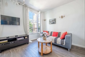 A living room with a flat-screen TV, a sofa with patterned cushions, a small round table with a vase, and a balcony with a view of buildings. - Bright & Central Duplex - Near the Eiffel Tower (Paris)
