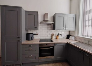A compact kitchen with grey cabinets, a built-in oven, a coffee maker, and a window with curtains. - Sweeting Street Apartment (Liverpool)