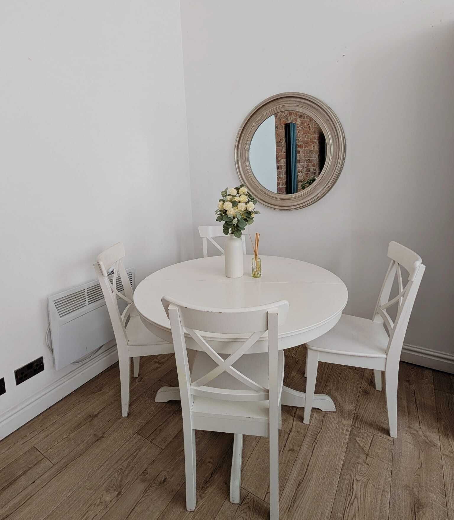 A round white table with four white chairs, a vase with flowers, and a mirror on the wall.