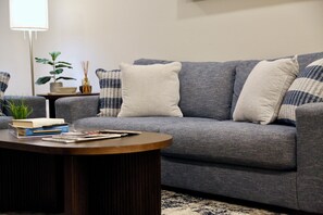 Un sofá gris con dos cojines blancos y azules estampados, una mesita auxiliar con una planta y una vela, y una mesa de centro redonda de madera con libros y revistas.