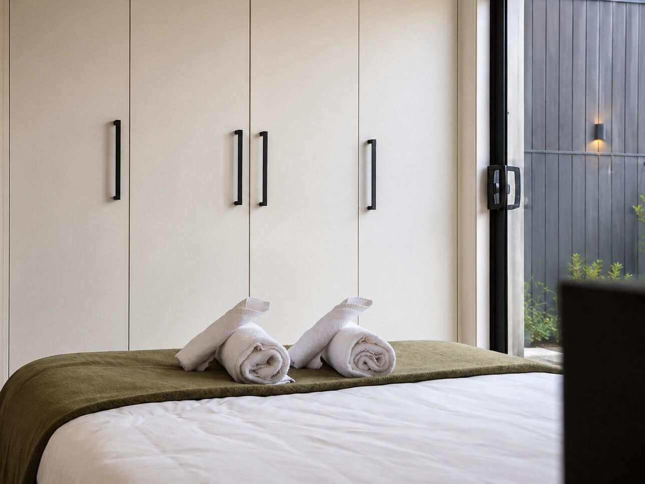 A hotel room with a bed, two white towels, and a window with a view of greenery.