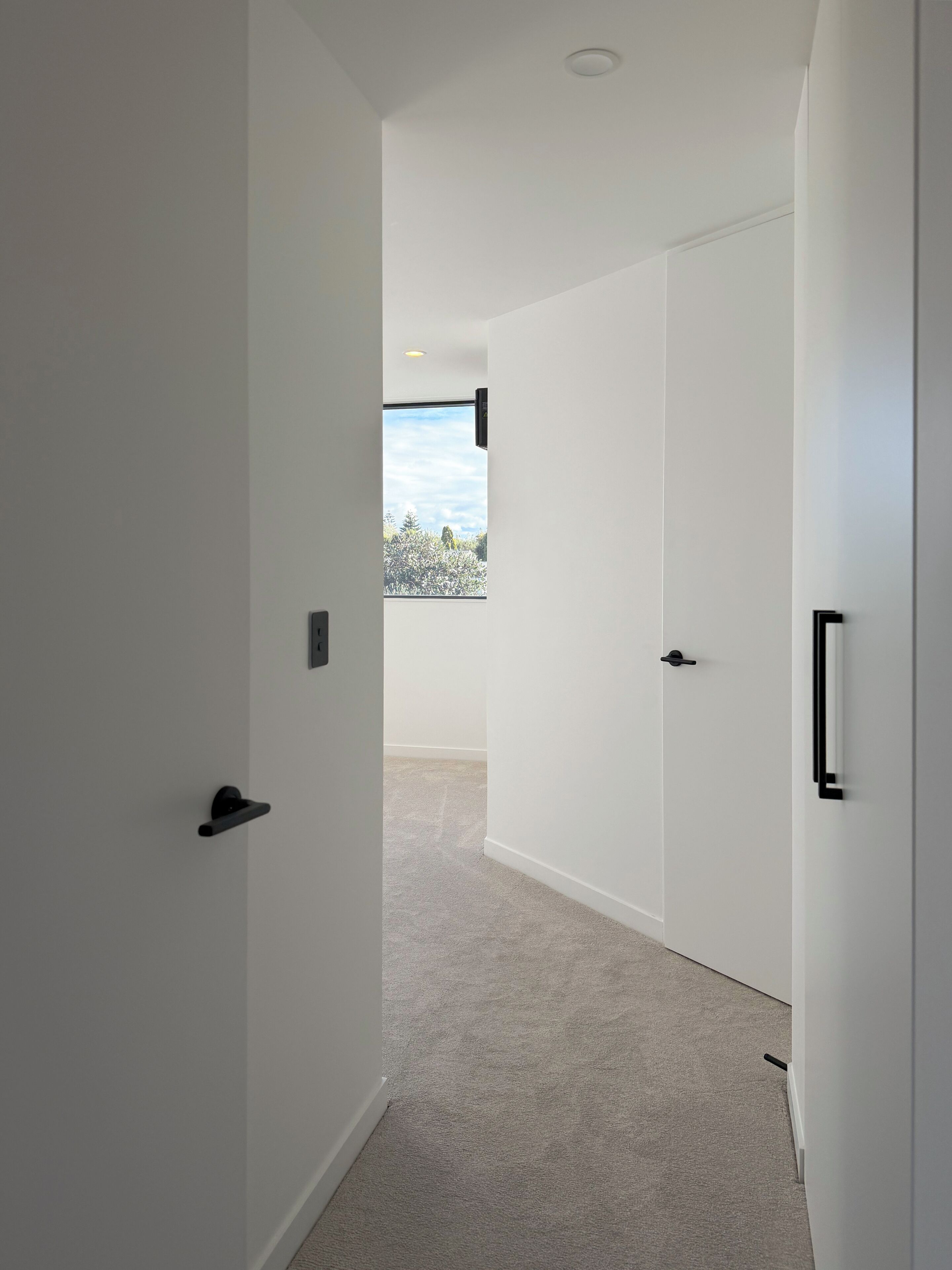 A hallway with white walls, a carpeted floor, and a door leading to a room with a window.
