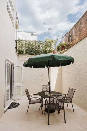 A patio with a table and chairs, an umbrella, and a white brick wall. - Stunning Garden Flat in Heart of Cheltenham (Cheltenham)