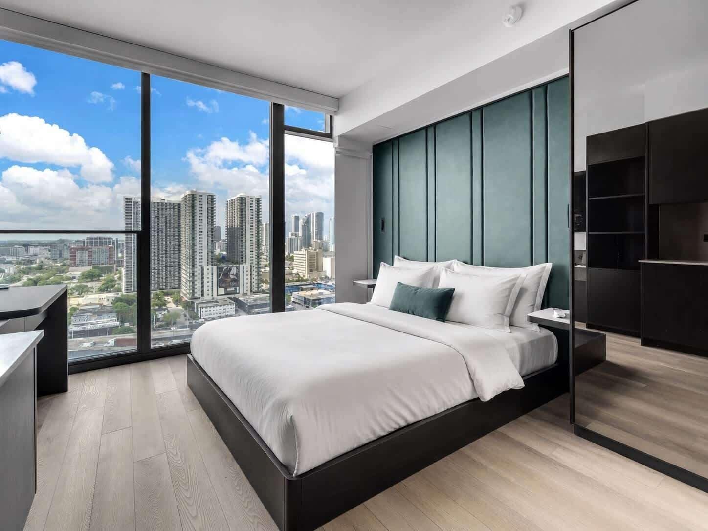 Ein modernes Schlafzimmer mit einem großen Bett, einem Schreibtisch und Blick auf eine Stadtlandschaft durch raumhohe Fenster.