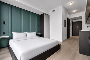 Ein modernes Hotelzimmer mit einem großen Bett, einem Nachttisch, einer eingebauten Sitzbank, einem Kleiderschrank, einem Badezimmer und einer Küchenzeile.
