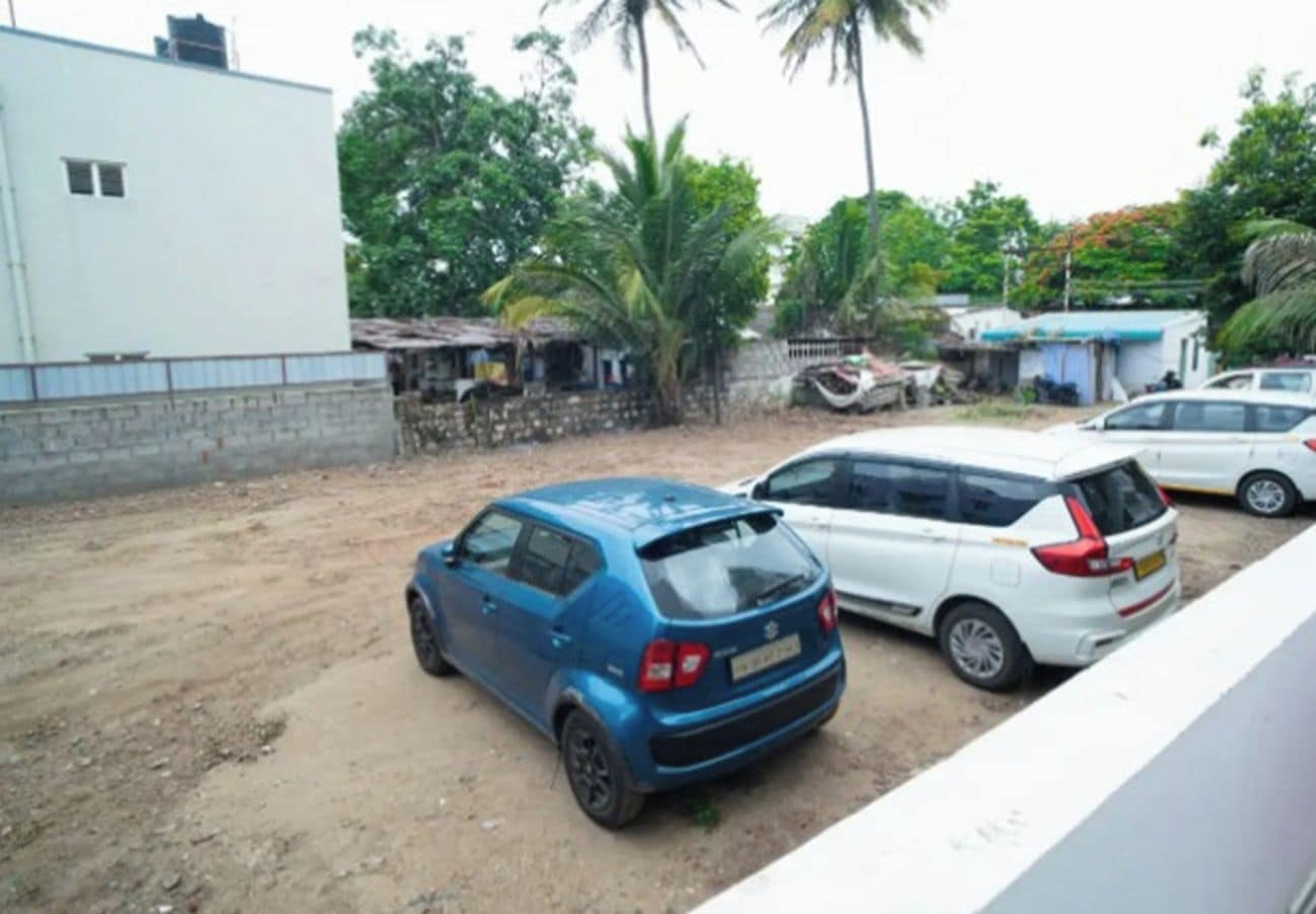 Un aparcamiento con un coche azul y una furgoneta blanca, rodeado de vegetación y un edificio.