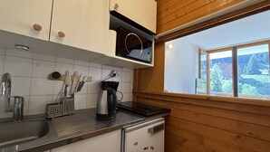 A kitchen with white cabinets, a black countertop, a coffee maker, and a window with a view of trees and buildings. - Studio apartment at the base of the ski lifts, in the center of the resort (Modane)