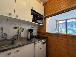 A kitchen with white cabinets, a black countertop, a coffee maker, and a window with a view of trees and buildings. - Studio apartment at the base of the ski lifts, in the center of the resort (Modane)
