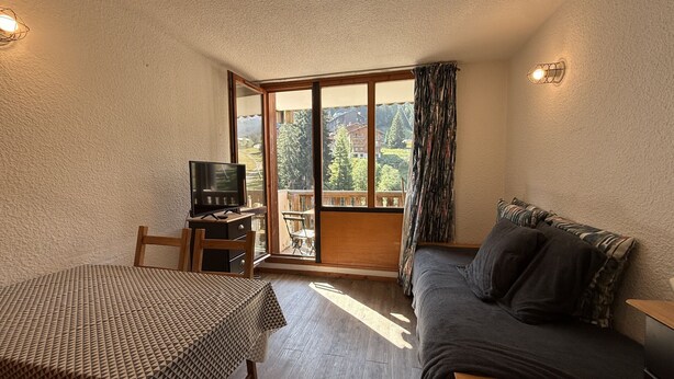 A room with a wooden table, a sofa, a television, and a sliding glass door leading to an outdoor area with a view of trees and buildings. - Studio apartment at the base of the ski lifts, in the center of the resort (Modane)