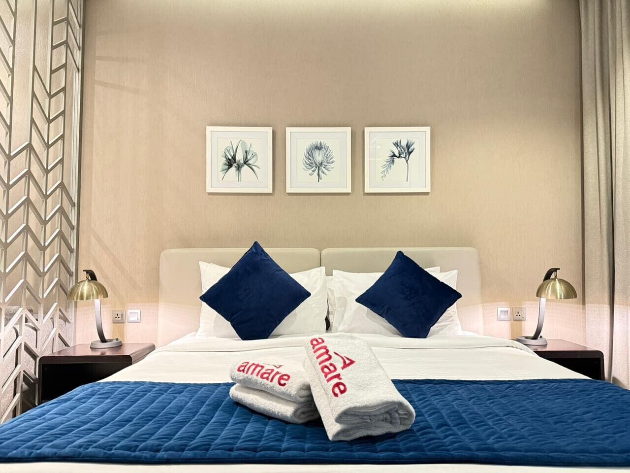Habitación de hotel con cama, almohadas, colcha azul acolchada, toalla doblada con la marca 'Amare' e impresiones botánicas enmarcadas en la pared.