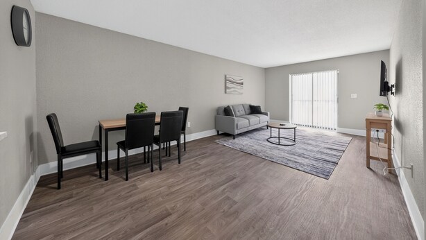A modern living room with a dining area, grey walls, wooden flooring, and a large window with blinds. - Stylish Furnished Retreat with Modern Comforts (Tamarac)