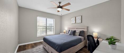 Un dormitorio con cama, escritorio, silla, ventilador de techo, ventana y una planta.