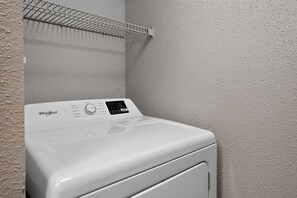 A Whirlpool front-loading washing machine in a laundry room with a metal shelf above it. - Cozy Contemporary Retreat with Great Amenities (Tamarac)