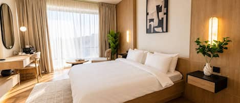 Una habitación de hotel con una cama grande, un escritorio, una silla, una mesita de noche con lámpara y un amplio ventanal con cortinas.
