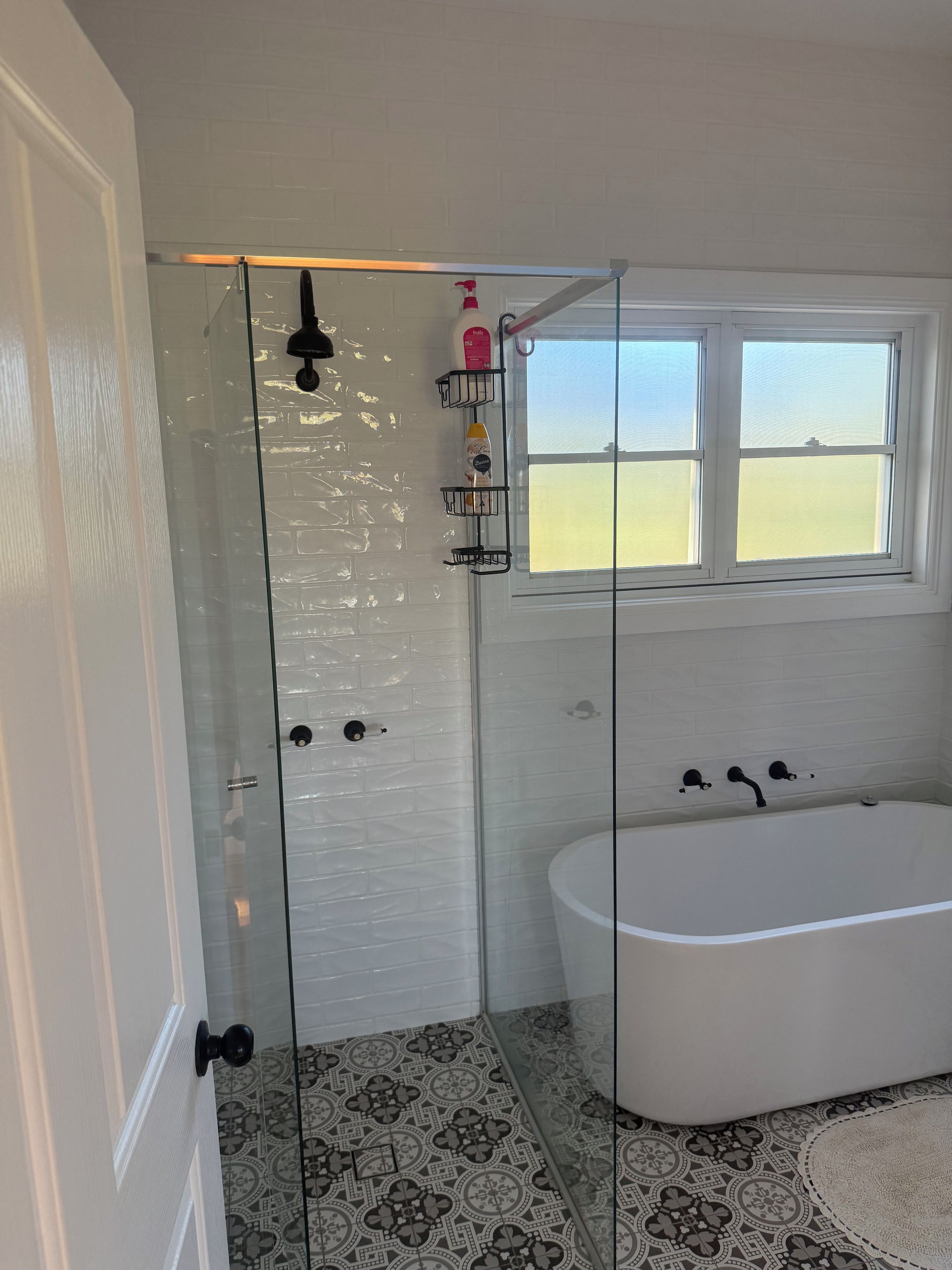 A bathroom with a glass shower enclosure, a white bathtub, patterned floor tiles, and a window with a view.