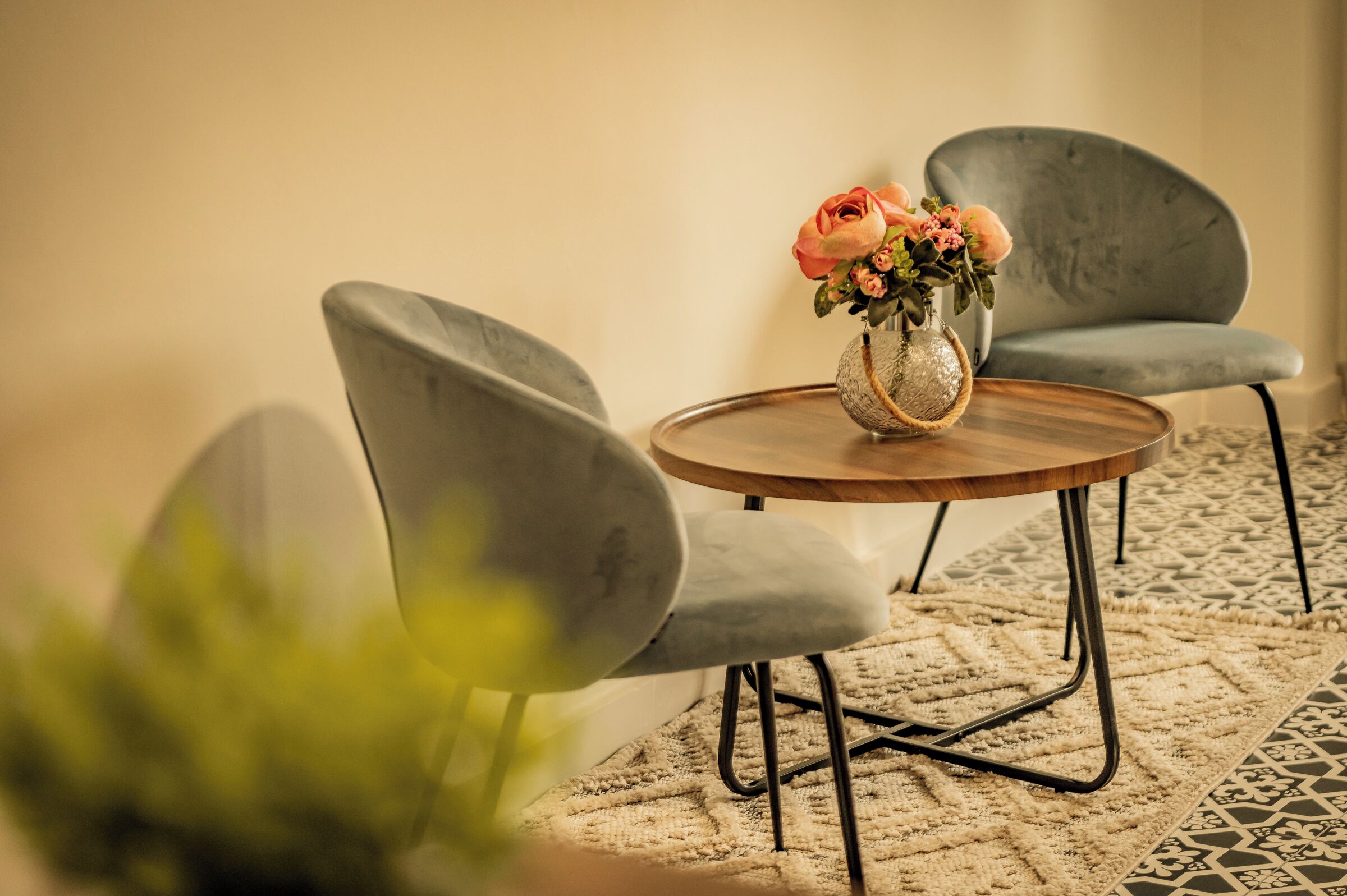 A round wooden table with a vase of flowers, two grey upholstered chairs, and a patterned rug.