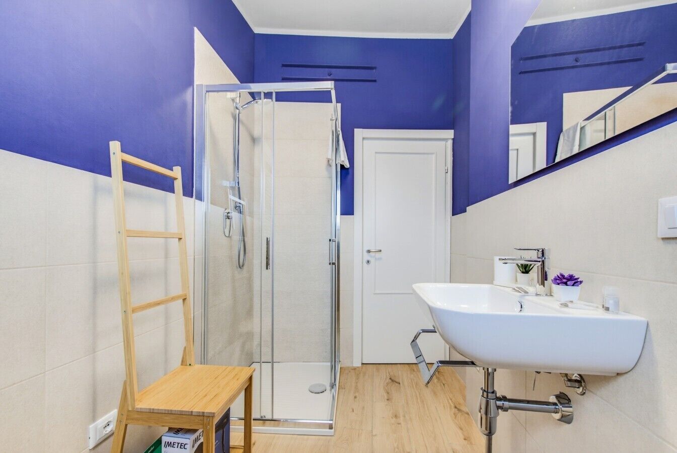 A bathroom with a glass shower enclosure, a white sink with a mirror, and a wooden stool.