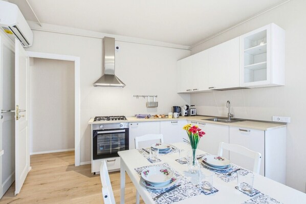Cocina compacta con armarios blancos, estufa a gas, horno y una mesa de comedor con platos y un jarrón de flores.