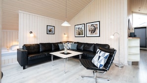 Ein modernes Wohnzimmer mit einem schwarzen Ledersofa, einem weißen Couchtisch und einem schwarzen Drehsessel. An der Wand hängen drei gerahmte Bilder, eine Stehlampe und ein großes Fenster mit Jalousien.