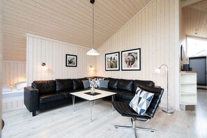 A modern living room with a black leather sectional sofa, a white coffee table, and a black swivel chair. There are three framed pictures on the wall, a floor lamp, and a large window with blinds. - Coastal Wellness House fur 10 Personen (Grossenbrode)