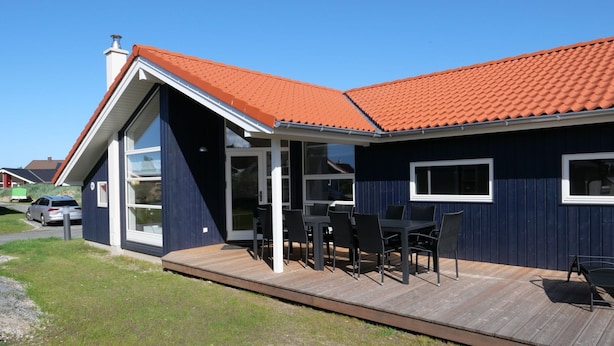 Ein modernes Haus mit rotem Ziegeldach, einer überdachten Terrasse mit Gartenmöbeln und einer großen Glasdoor, die auf eine Holzterrasse führt.