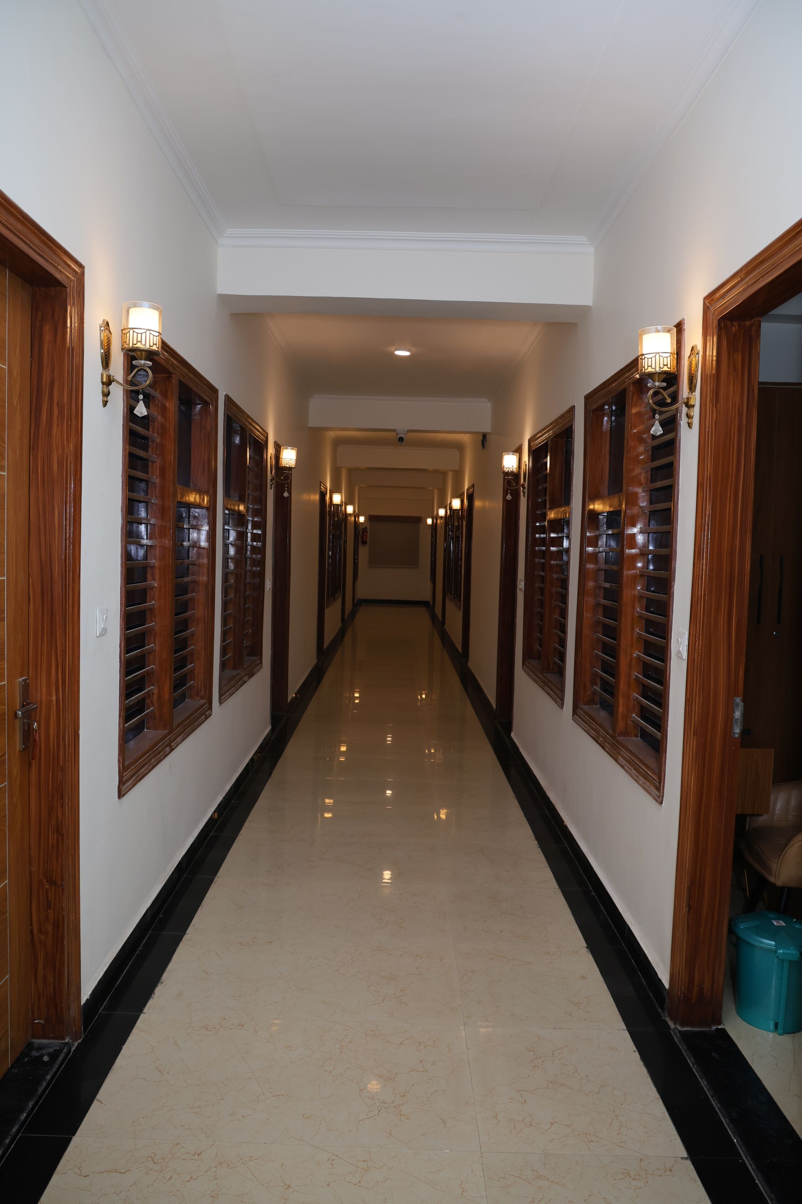 Um corredor longo e estreito, com portas de madeira e arandelas na parede, que leva a um quarto com ventoinha no teto.