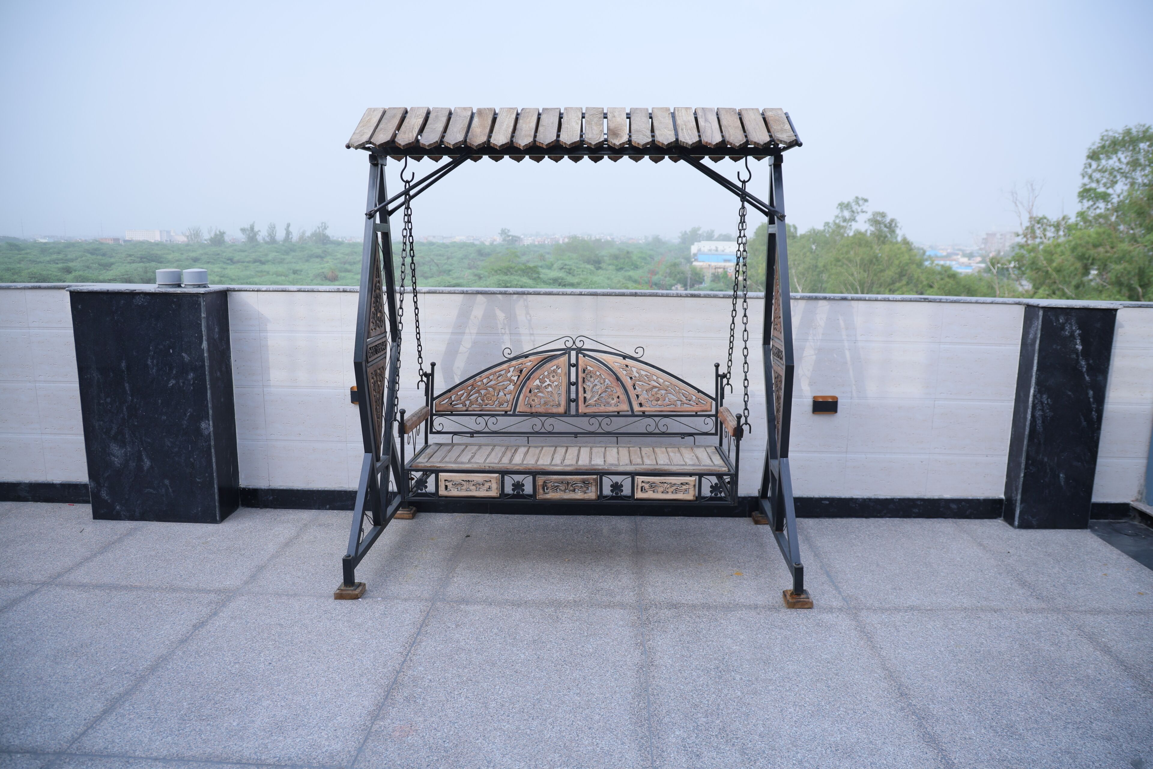 Una hamaca con asiento de madera y estructura metálica, ubicada en una terraza en la azotea con vistas a zonas verdes y edificios a lo lejos.