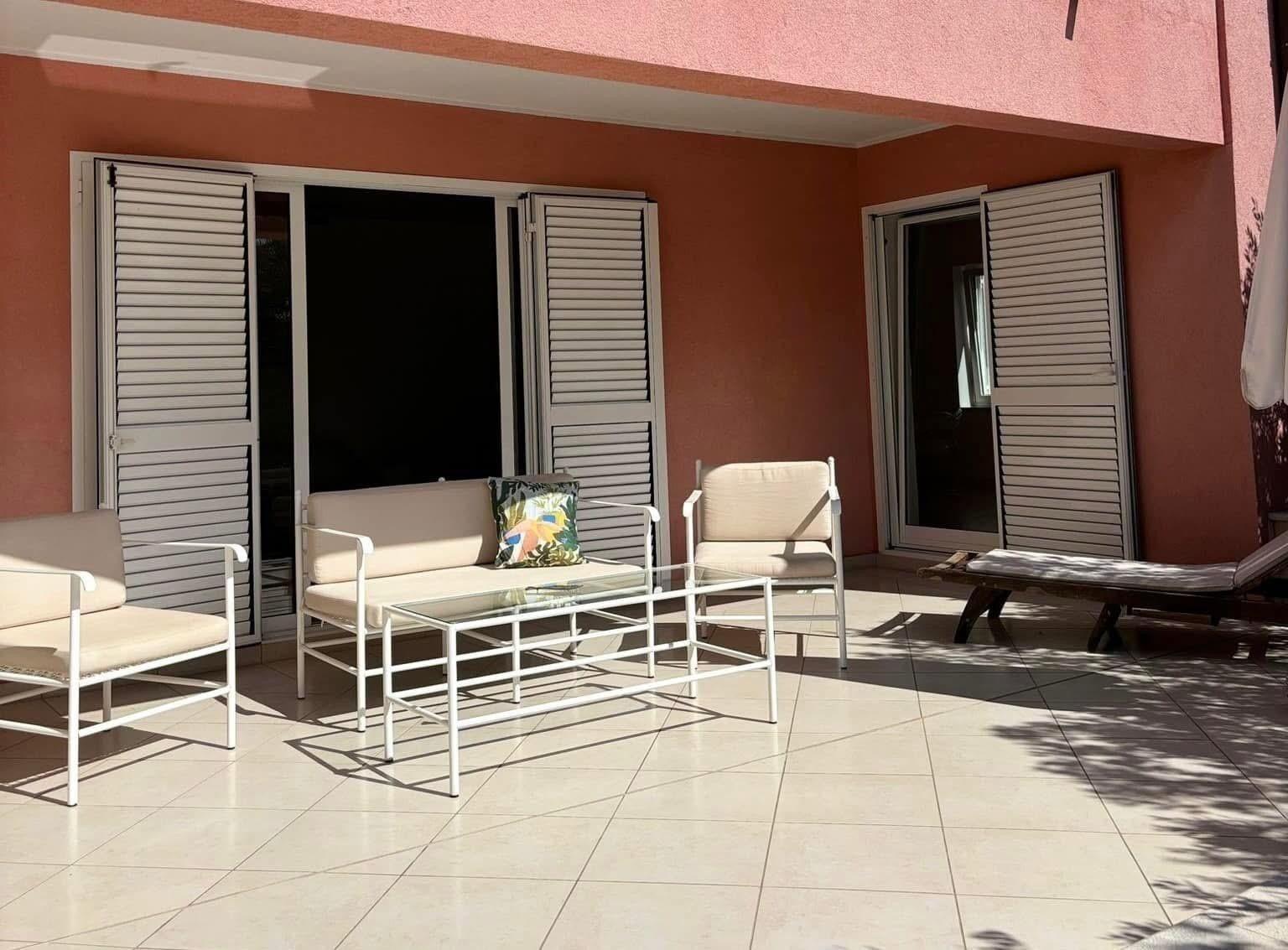 Ein Sitzbereich im Freien mit einer Couch, Stühlen und einem Tisch, umgeben von großen Fenstern mit Fensterläden.
