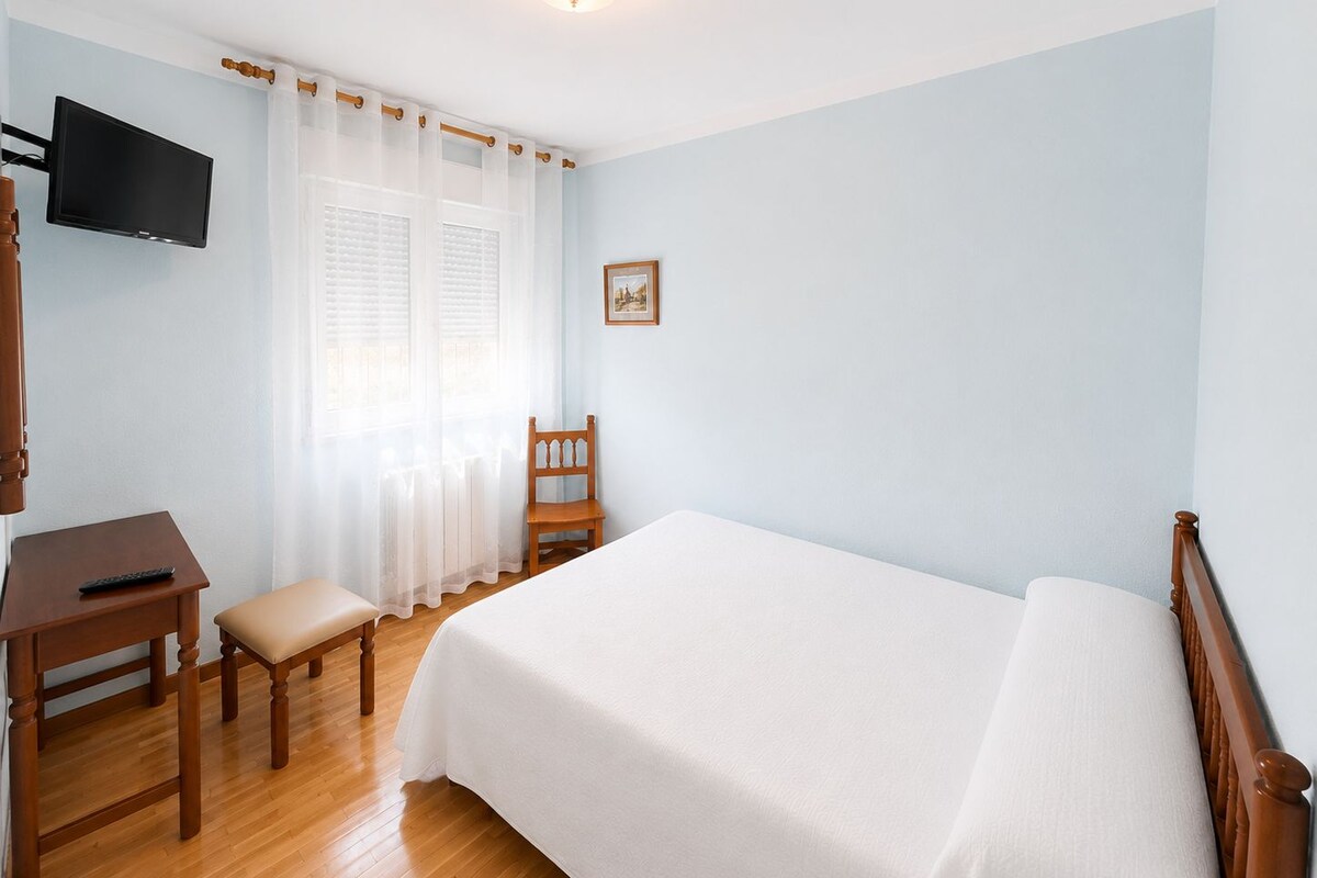 Un dormitorio con cama, escritorio, silla, televisor y un cuadro en la pared.