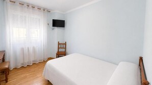 Una habitación con cama, una silla, un televisor montado en la pared y un ventanal con cortinas.