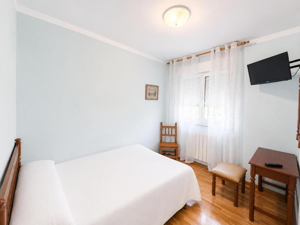 Ein Schlafzimmer mit einem Bett, einem Sessel, einem Schreibtisch, einem Fernseher und einem Fenster mit Vorhängen.