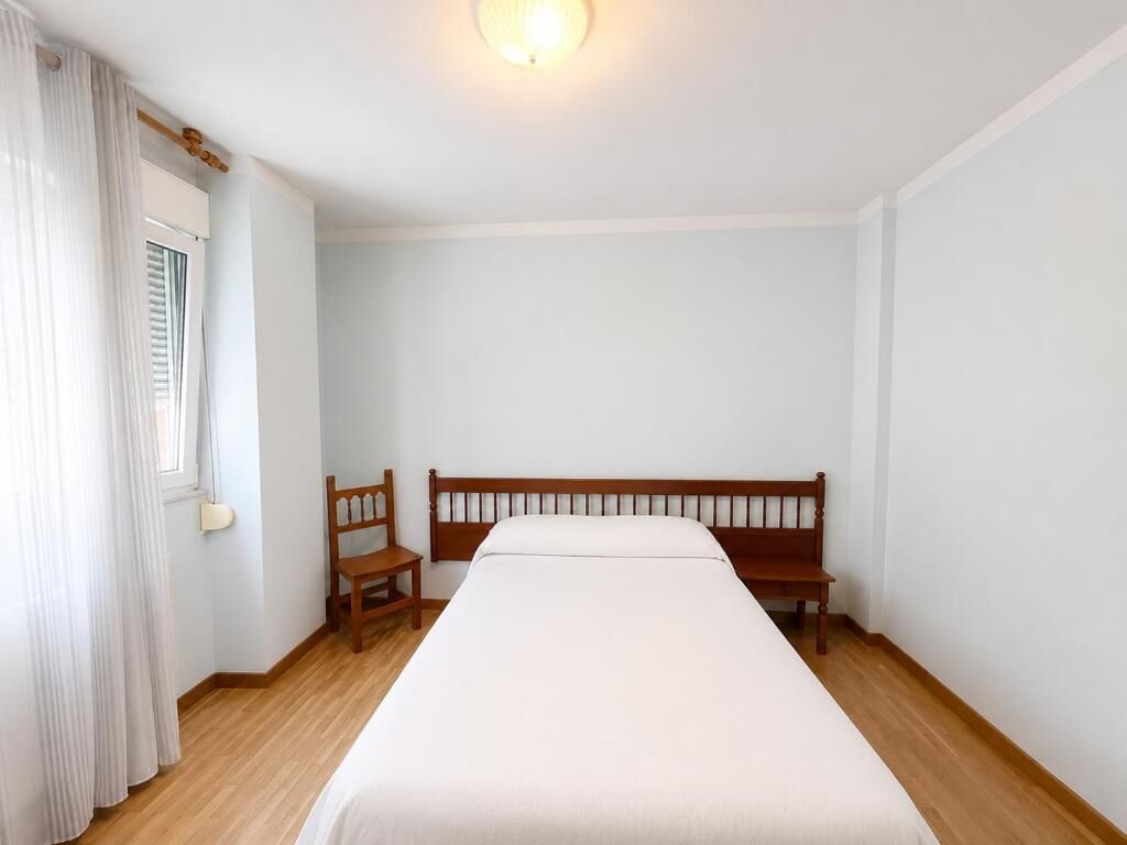 Una habitación con una cama individual, una silla de madera, una ventana con cortinas y una lámpara fijada a la pared.