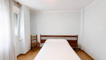 Una habitación con una cama individual, una silla de madera, una ventana con cortinas y una lámpara fijada a la pared.