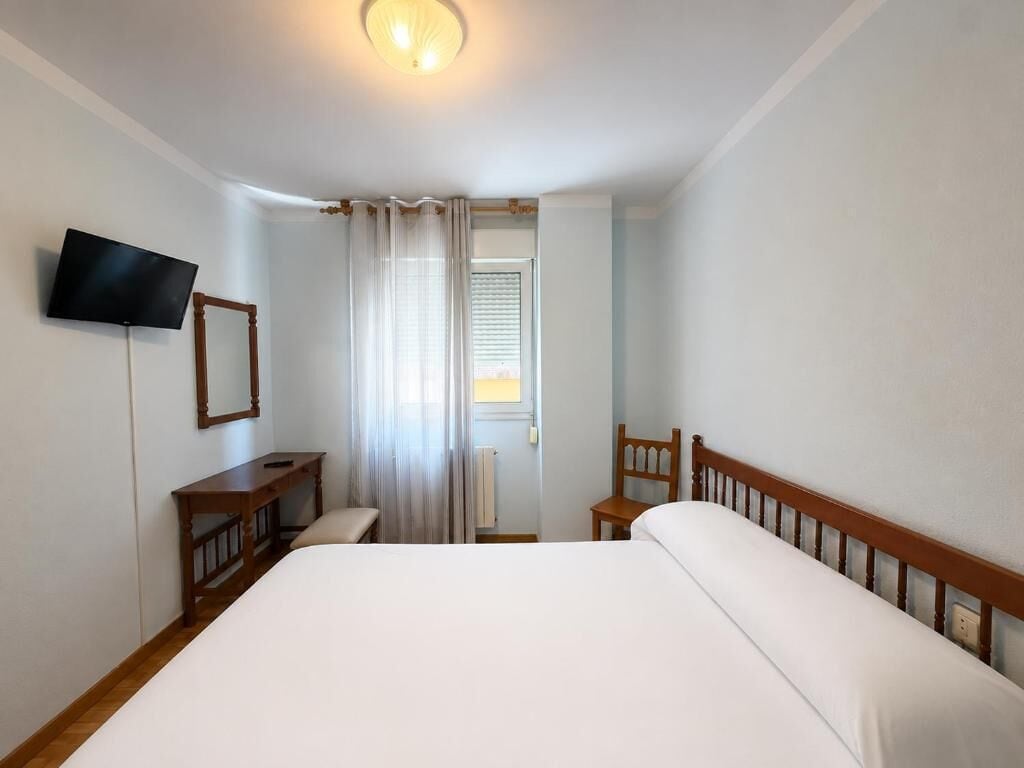 Een hotelkamer met een bed, een aan de muur bevestigde televisie, een houten stoel, een klein tafeltje en een raam met gordijnen.