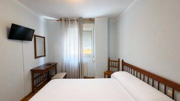 Een hotelkamer met een bed, een aan de muur bevestigde televisie, een houten stoel, een klein tafeltje en een raam met gordijnen.