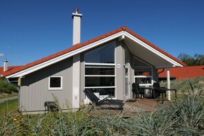 Ett modernt hus med ett rött tak, ett stort glasfönster och en terrass med utemöbler.