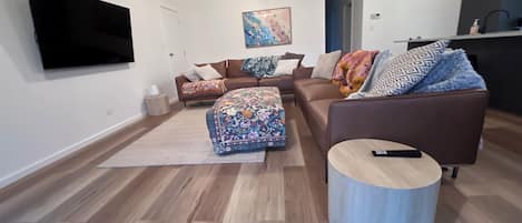A modern living room with a flat-screen TV, a sofa set, an ottoman, and a round coffee table.