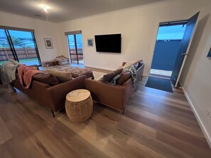 A modern living room with a brown leather sofa, a round wooden side table, and a flat-screen TV mounted on the wall.
