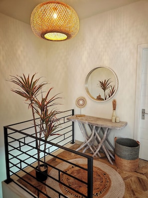 Eine Treppe mit schwarzer Metallreling, ein gewobener Rattanwandbehang, ein runder Spiegel mit Zierrahmen und ein Holztischchen mit Dekoration.