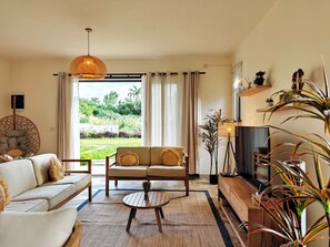 A living room with a sofa, a coffee table, and a television. There is a window with curtains, a hanging light fixture, and a plant. - Designer Luxury villa, 10 mins walk from beach (Grand Baie)