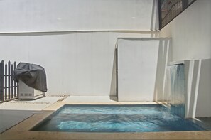 Une piscine à l’eau d’un bleu limpide, entourée d’un mur blanc et d’un barbecue couvert.