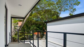 En smal balkon med gelænder i metal, der fører til et overdækket udend ørsområde med træer og en bygning i baggrunden.