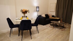 Uma área de refeições com uma mesa redonda, duas cadeiras e um vaso com flores. Há um sofá e uma mesa de centro ao fundo.