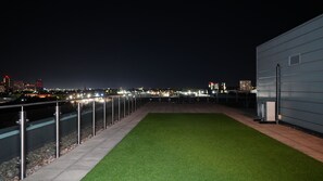 A rooftop terrace with a view of a cityscape at night, a metal railing, and a grassy area. - Luxury 2-Bed Apartment | 3 Bathrooms | Balcony & Rooftop Terrace (Salford)