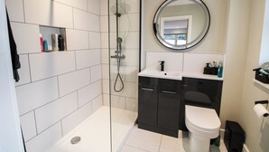 A modern bathroom with a glass-enclosed shower, a round mirror, a white toilet, and a dark vanity with a sink. - Cefn Coed (Presteigne)